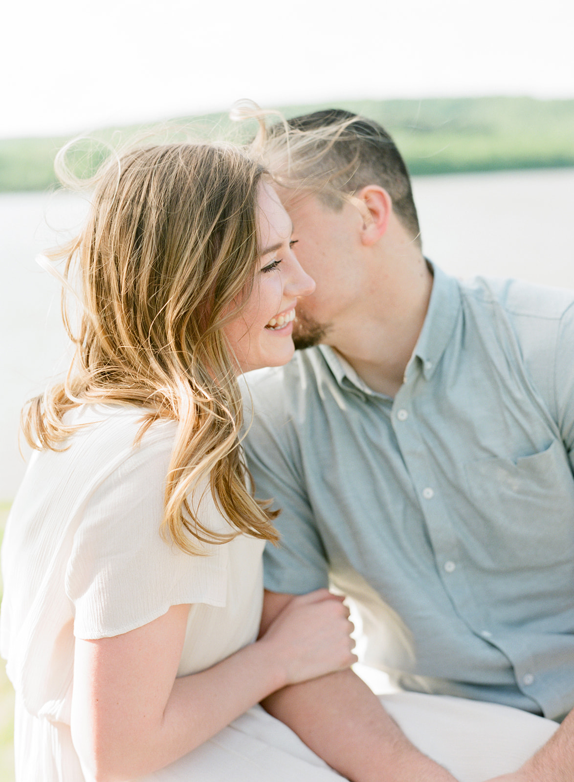 engagement session at okmulgee lake in oklahoma with girl laughing off into the distance while guy whispers into her ear and the sun in shining on their back